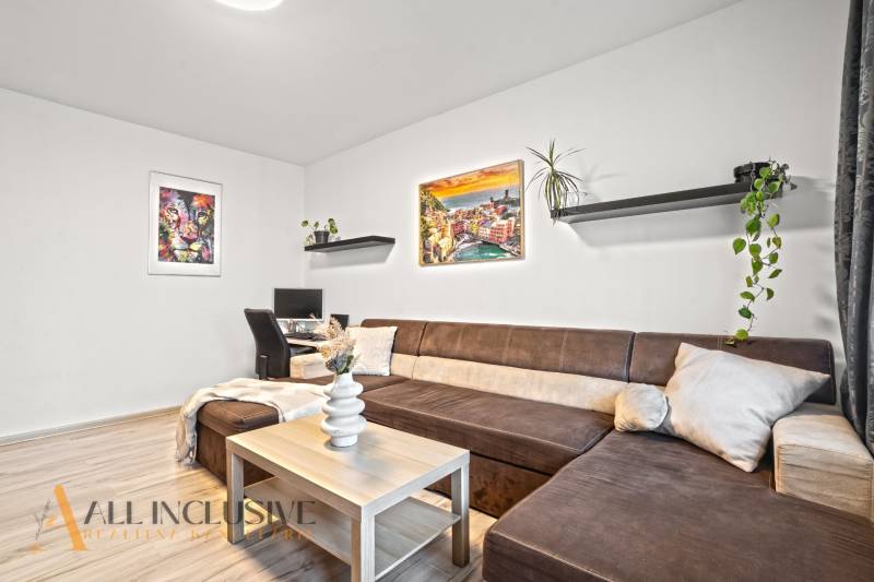 NA PREDAJ 3-IZBOVÝ BYT S PIVNICOU, SENEC - Obývacia izba s hnedou sedačkou, pracovným stolom a podlahou s dreveným dekorom.