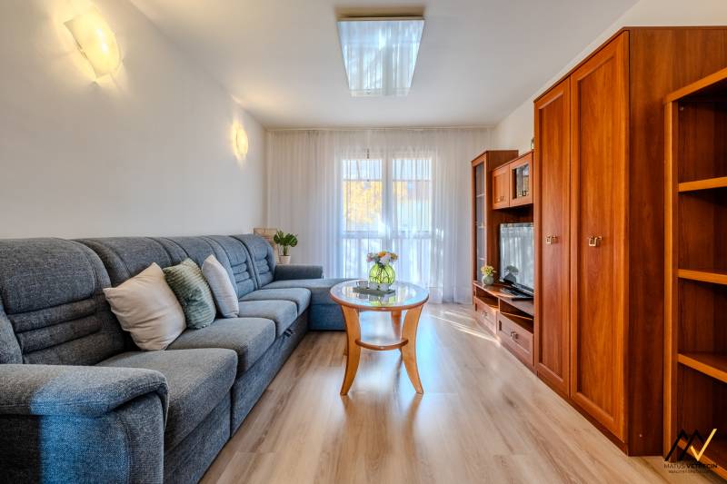 2 IZBOVÝ BYT PREŠOV, ULICA TARASA ŠEVČENKA - Obývacia izba v 2-izbovom byte s podlahou s dreveným dekorom, pohodlnou sedačkou a nábytkovou stenou.