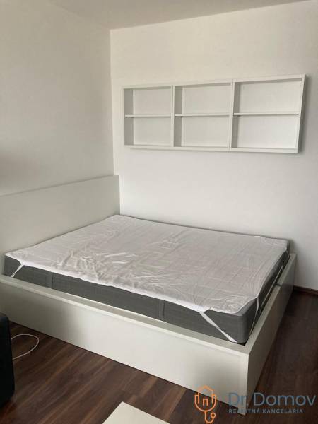 NA PRENÁJOM ÚTULNÝ 1-IZBOVÝ BYT, ČIASTOČNÁ REK., KAROLA ADLERA, BA - Spálňa v 1-izbovom byte s posteľou, bielymi policami a podlahou s dreveným dekorom.