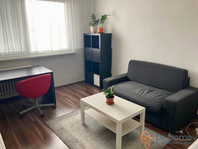 NA PRENÁJOM ÚTULNÝ 1-IZBOVÝ BYT, ČIASTOČNÁ REK., KAROLA ADLERA, BA - 1-izbový byt s pohovkou, stolom, červenou stoličkou, poličkou a podlahou s dreveným dekorom.