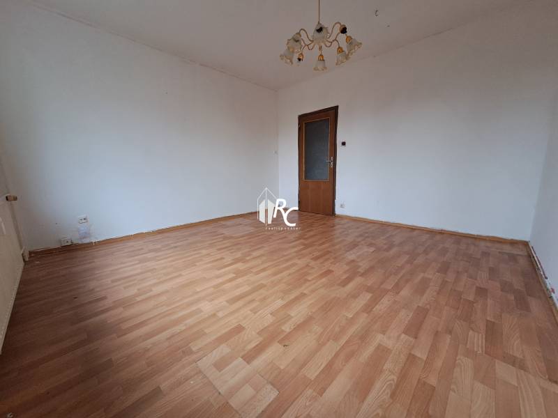 REZERVOVANÉ 2-izbový byt s nádherným výhľadom v pôvodnom stave   - Izba v 2-izbovom byte s podlahou s dreveným dekorom a lustrom.