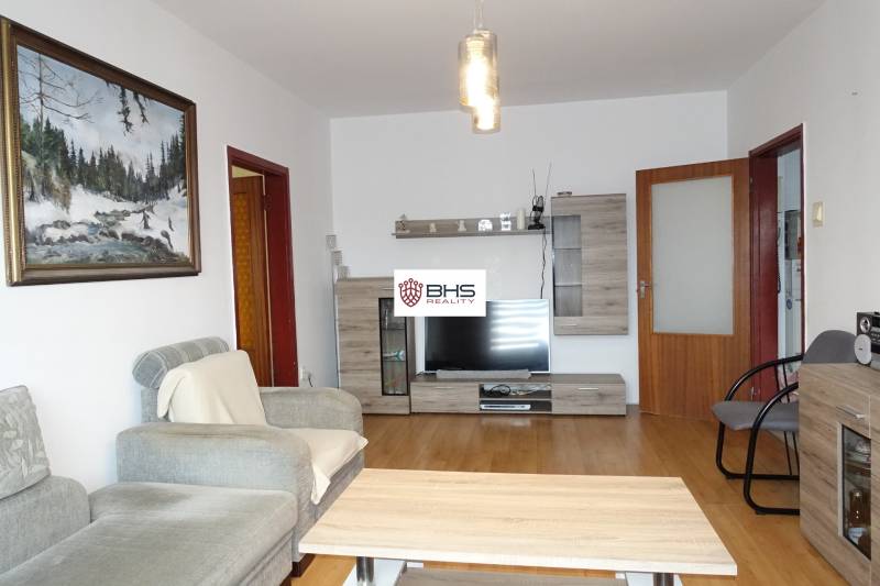 PREDAJ- 3-iz. byt, Homolova - Dúbravka, 67 m2, 11./12. posch., lodžia - Obývačka 3-izbového bytu s pohodlnou pohovkou, nábytok s dreveným dekorom a televízorom.