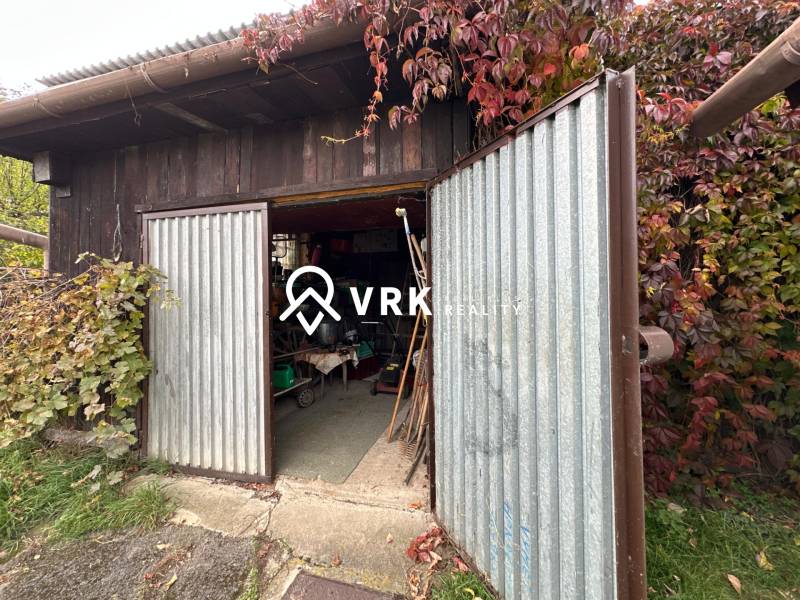 RODINNÝ DOM - CHALUPA, KALŠA - Otvorené kovové dvere garáže pri rodinnom dome v Kalši, obklopené bujnou vegetáciou.