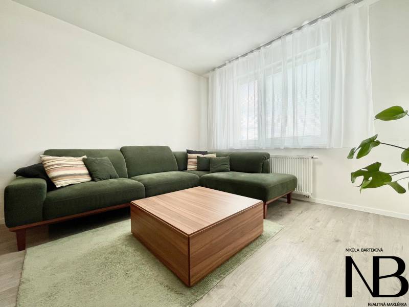 Na predaj štýlový 2-izbový byt s loggiou a parkovaním – TN, Na Pažiti - Obývacia izba s pohodlnou zelenou sedačkou, stolíkom s dreveným dekorom a veľkým oknom.
