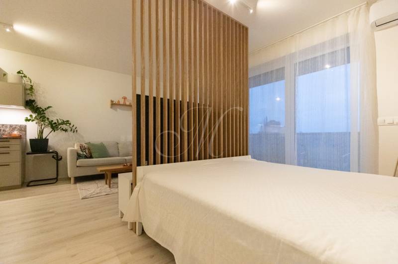 Slnečný 1,5i byt s loggiou a parkovaním - Kuchyňa so svetlým nábytkom, pohovkou a lôžkom v 1-izbovom byte, podlaha s dreveným dekorom.