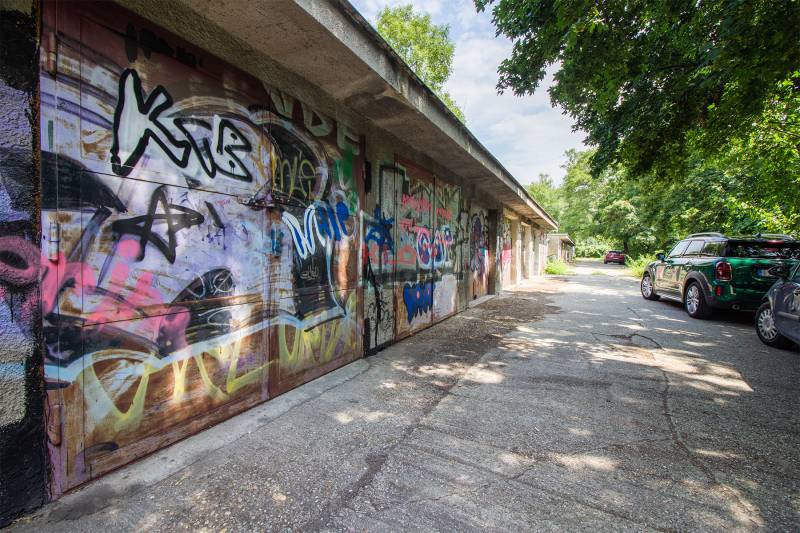 Pozrite si 17m², ZREKONŠTRUOVANÚ garáž, pri nemocnici Kramáre - Graffiti pokrývajú garáže na ulici Opavská, autá parkujú na ceste obklopenej stromami.