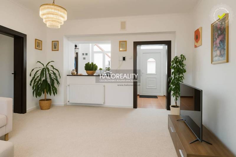 Predaj, rodinný dom Beša - Obývacia izba v rodinnom dome s bielymi stenami, kvetinami a televízorom na stolíku.