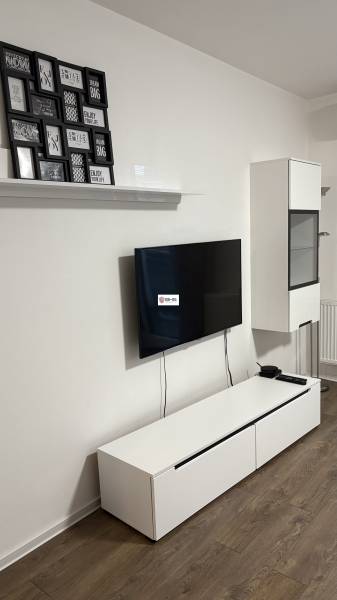 Predaj 1ibyt (garsonka) v uzavretom areáli v DNV (novostavba) - Televízor na stene, biela komoda, obrazy a podlaha s dreveným dekorom v 1-izbovom byte.
