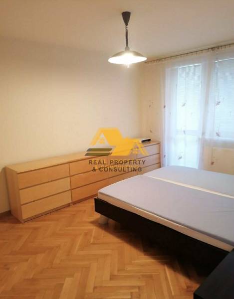 Prenajmem 3 izbový byt v Nových Zámkoch - Spálňa v 3-izbovom byte s posteľou a komodou na podlahe s dreveným dekorom.