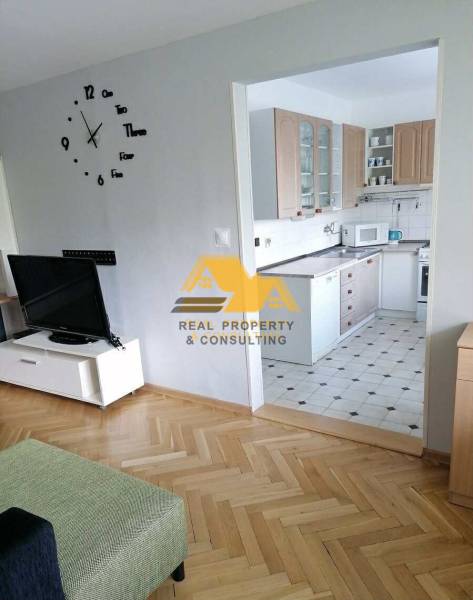 Prenajmem 3 izbový byt v Nových Zámkoch - Obývačka v 3-izbovom byte s podlahou s dreveným dekorom a kuchyňou v pozadí.