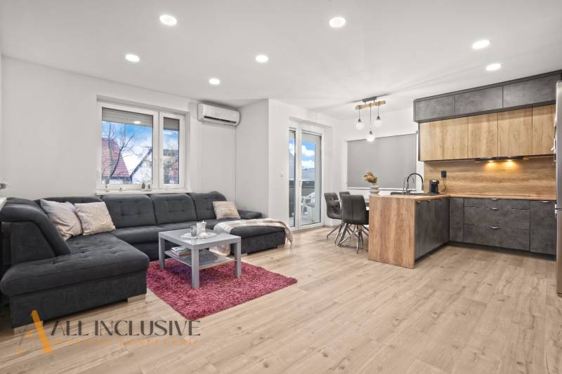 REZERVOVANÉ | NA PREDAJ 3-IZBOVÝ BYT S BALKÓNOM, VRAKÚŇ - Obývačka s kuchynským kútom, šedá sedačka, podlaha s dreveným dekorom, jedálenský stôl.