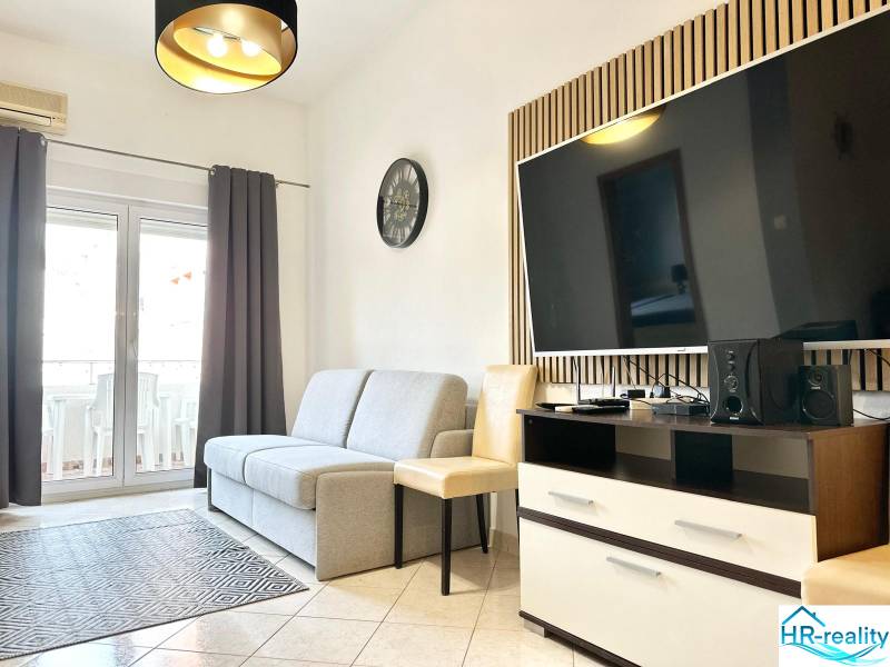 Trogir, Čiovo – pekný, zariadený apartmán len 150m od pláže - Obývacia izba s pohovkou a televízorom v rekreačnom apartmáne, svetlá dlažba, balkónové dvere.