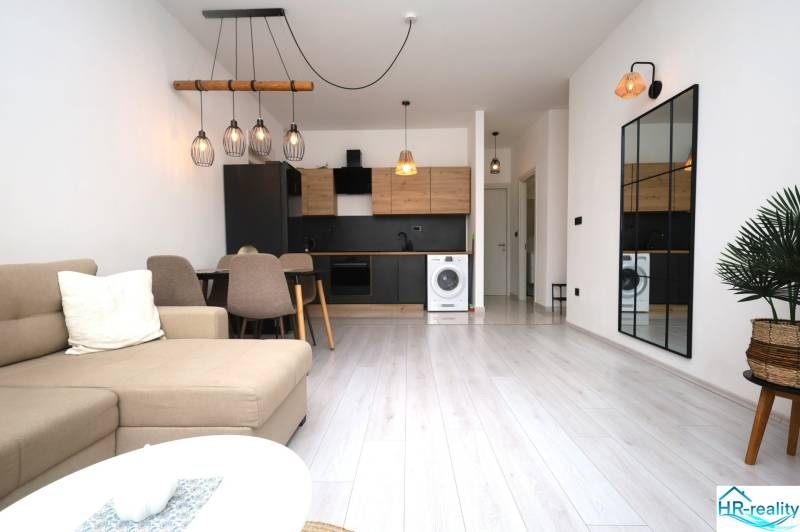 Vodice – novo zrekonštruovaný dvojspálňový apartmán s výhľadom na more - Obývacia izba Rekreačného apartmánu s kuchyňou, stolom, zrkadlom a podlahou s dreveným dekorom.