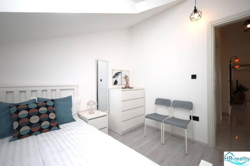 Vodice – novo zrekonštruovaný dvojspálňový apartmán s výhľadom na more - Rekreačný apartmán so svetlou spálňou, posteľou, komodou a vankúšmi, podlaha s dreveným dekorom.