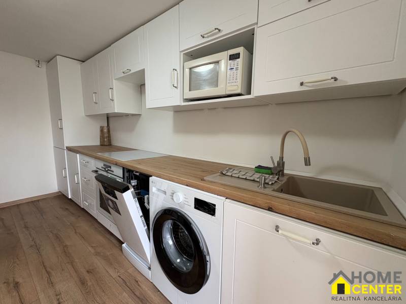 NA PRENÁJOM krásny 1 izbový apartmán so záhradou v srdci Komárna - Kuchyňa v 1-izbovom byte s bielou linkou, práčkou a podlahou s dreveným dekorom.