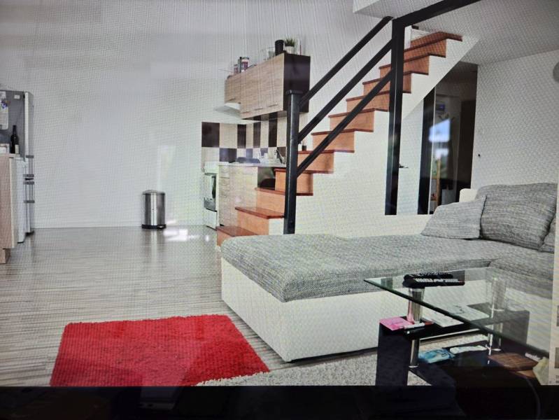 Na predaj ubytovňa / rodinný dom  v Šali  Veči s kapacitou 50 osôb - Obývacia izba s kuchynským kútom, schodiskom, gaučom, skleneným stolíkom a podlahou s dreveným dekorom.