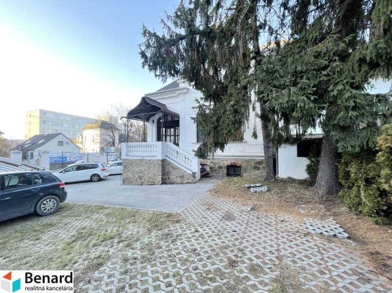 PREŠOV-ŠIRŠIE CENTRUM,POLYFUNKČNÁ BUDOVA,VL.PARKOVISKO,UL.VAJANSKÉHO - Budova na Vajanského v Prešove s prístupovou cestou, autami a smrekmi v popredí.