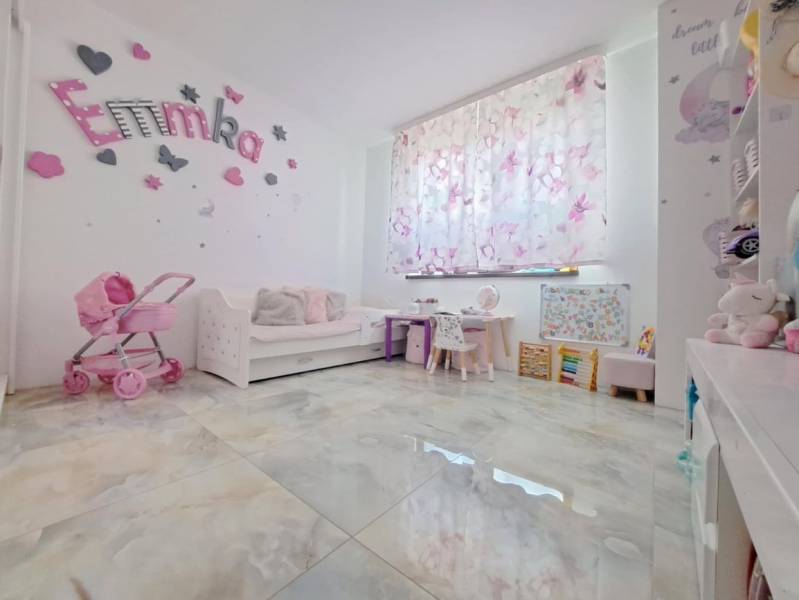 Neodolateľná ponuka! 4-izbová novostavba Galanta-Kolónia za super cenu - Detská izba v rodinnom dome s dekoráciami, kočíkom a písacími stolíkmi. Podlaha má drevený dekor.