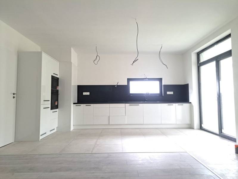 Priestranné novostavby 4-izbových rodinných domov v meste Šaľa - Kuchyňa v rodinnom dome s podlahou s dreveným dekorom, bielymi skrinkami a vstavanými spotrebičmi.