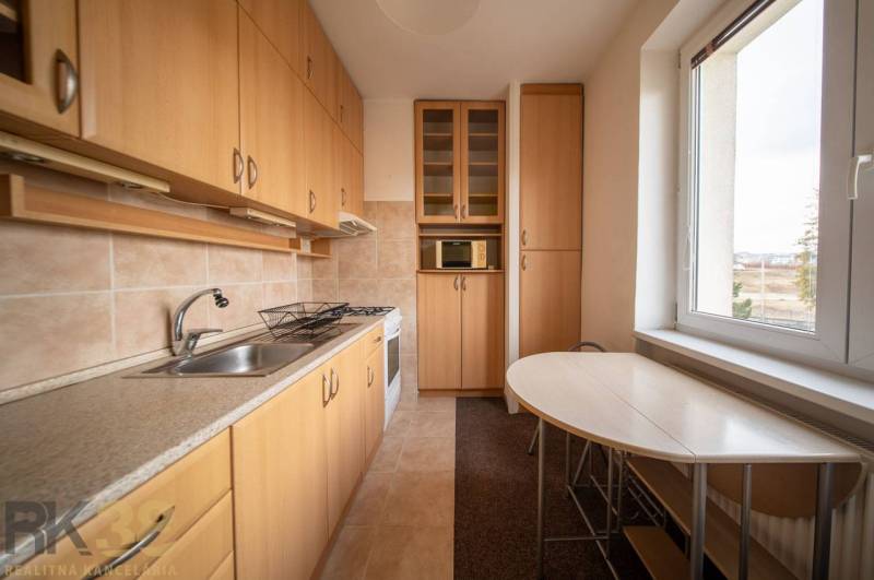 Rezervované: Na predaj pekný 2-izbový byt v centre mesta Poprad - Kuchyňa v 2-izbovom byte s podlahou s dreveným dekorom, svetlým nábytkom a stolom pri okne.