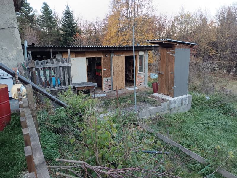 Samota Nová Baňa – Štále s hospodárskymi stavbami  - Drevený záhradný prístrešok s trávou, vedľa rodinného domu, obklopený stromami a záhradným náčiním.