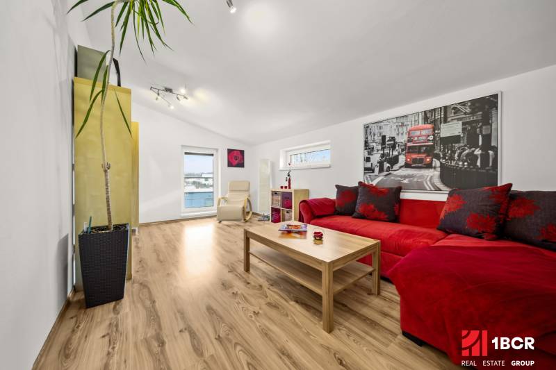 MODERNÝ 5-IZBOVÝ RODINNÝ DOM V ČUNOVE: 180m2 KOMFORTU A TICHA - Obývacia izba s podlahou s dreveným dekorom, červenou sedačkou a veľkou rastlinou.