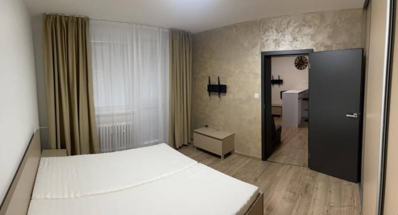 REZERVOVANÉ! Nadštandardný 1-izbový byt, ul. 1. mája, Vráble - Spálňový priestor s posteľou, závesmi a podlahou s dreveným dekorom v 1-izbovom byte.