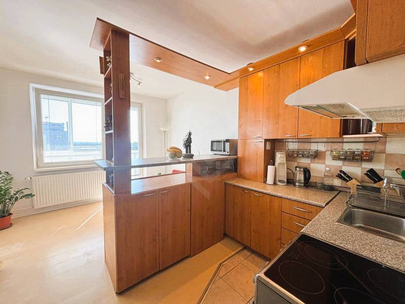 Slnečný dvojizbový byt – 43,5 m² v Bratislave – Vajnory - Kuchyňa v 2-izbovom byte s podlahou s dreveným dekorom, priestranným oknom a izbovou rastlinou.