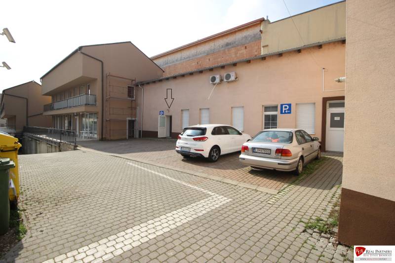 Ponúka na predaj budovu - centrum Senca - NOVÁ SUPER CENA  - Na ulici Mierové nám. pred budovou sú dve autá na parkovisku a smetné koše.