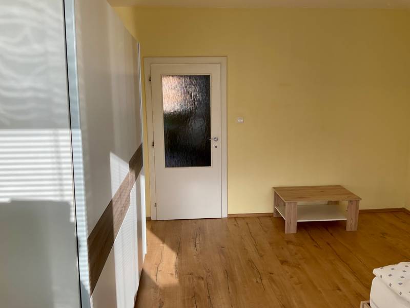 I prenájom štýlových izieb v 3-izbovom byte – Karlova Ves - Izba s podlahou s dreveným dekorom, svetlými stenami a jednoduchým nábytkom v 3-izbovom byte.