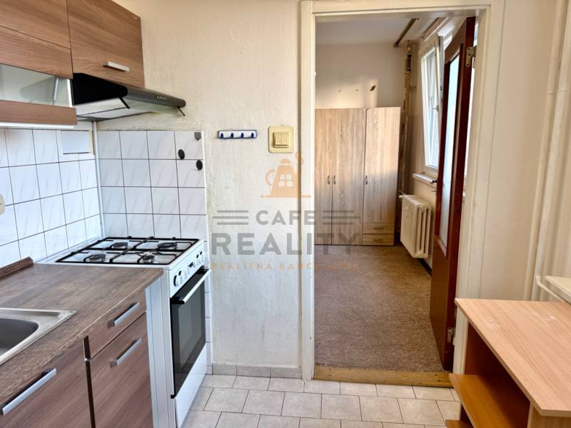 REZERVOVANÉ Na predaj 1 izbový byt, Bratislava, Dúbravka, Drobného - Kuchyňa s bielymi obkladačkami, sporákom, skrinkami a pohľad do predsiene v 1-izbovom byte.