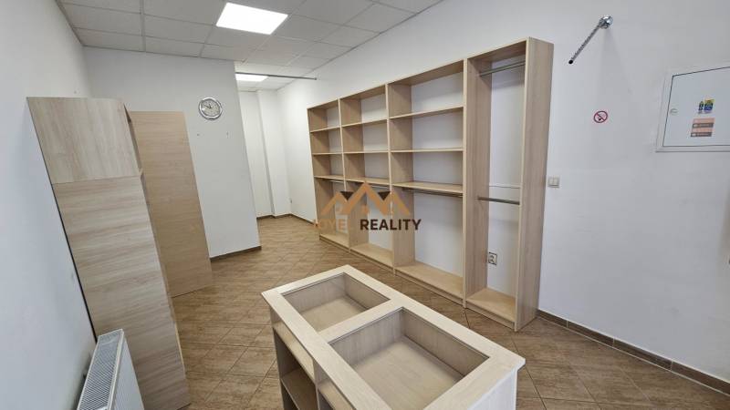 Na prenájom: Nebytový-obchodný priestor – Budovateľská ulica, Šaľa - Obchodné priestory s regálmi a podlahou s dreveným dekorom, biela stena, stropné osvetlenie.