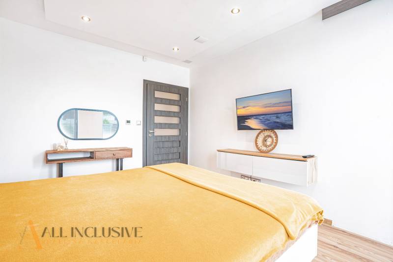 NA PREDAJ NADŠTANDARDNÁ 4-IZB. NOVOSTAVBA, DUN. KLÁTOV - Svetlá spálňa v rodinnom dome s televízorom, zrkadlom a žltou prikrývkou na posteli.