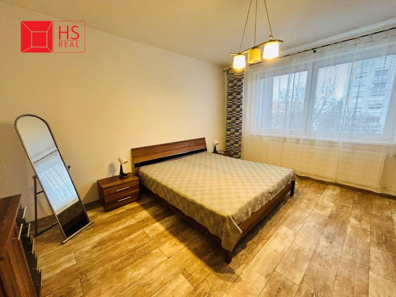 Prenajom 3 izb. bytu na Vajanskeho v NZ - Spálňa so zrkadlom, posteľou, svietidlom a podlahou s dreveným dekorom v 3-izbovom byte.