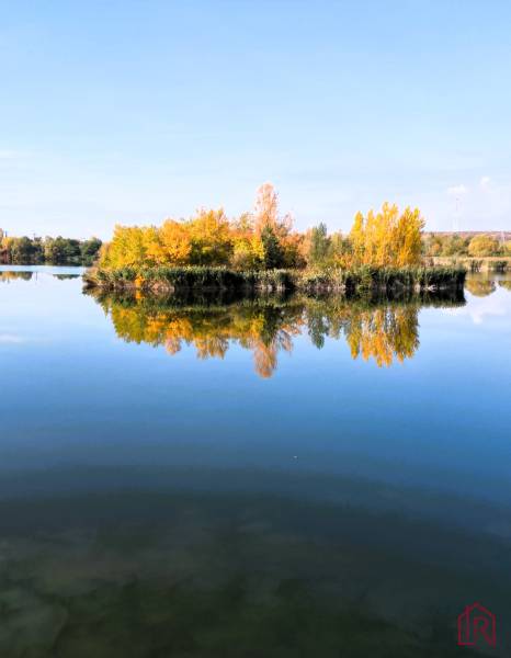 KH-1018, PREDAJ Pozemok pri jazere 887 m², garáž, altánok - Pokojná vodná hladina s jesennými stromami v Čani, odrážajúca výstavbu domov.