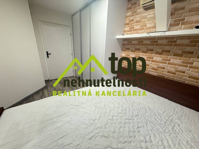 TOPOĽČANY Priestranný 1-izbový byt na prenájom 60 m2 - Spálňa s tehlovým obkladom steny a posteľou v 1-izbovom byte, podlaha s dreveným dekorom.