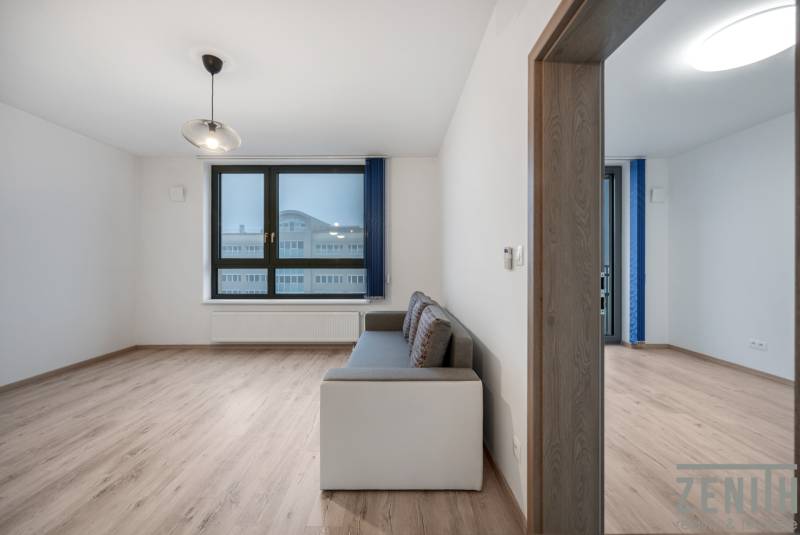 PREDAJ 2-IZBOVÉHO BYTU – NOVOSTAVBA JARABINKY, RUŽINOV - Obývacia izba so sedačkou, veľkým oknom a podlahou s dreveným dekorom v 2-izbovom byte.