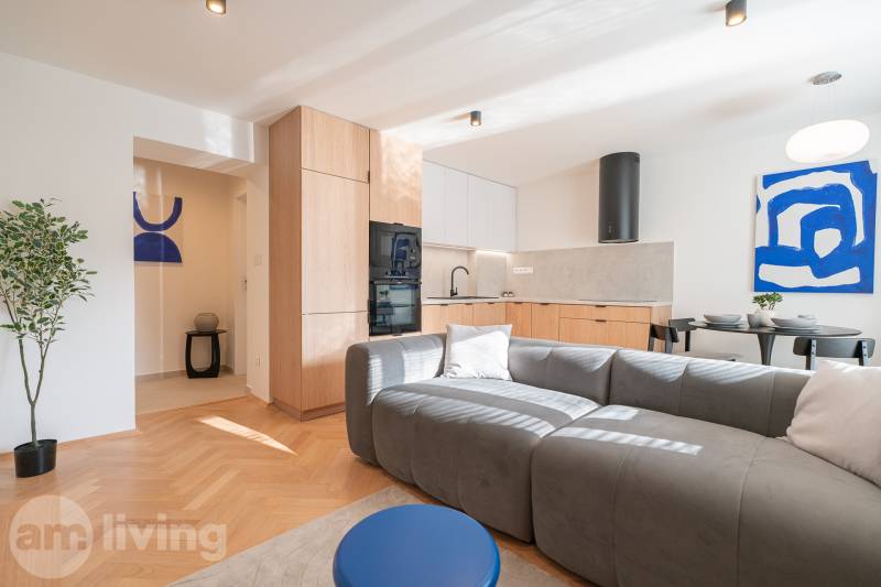 3i BYT V TOP LOKALITE – Trenčín, Sihoť - NA PRENÁJOM - Obývacia časť 3-izbového bytu s kuchyňou, sivou pohovkou a podlahou s dreveným dekorom.