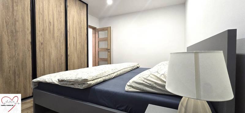 Prenájom komfortný, zariadený 3 izbový byt po úplnej rekonštrukcii - Spálňa s posteľou, skriňou s dreveným dekorom a lampou v 3-izbovom byte.