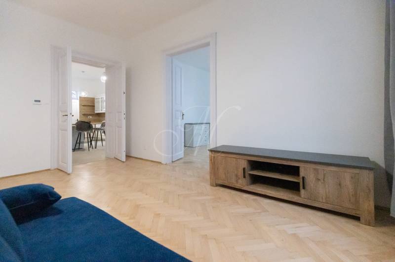 Priestranný 3i byt s vysokými stropmi, Dunajská - Obývacia izba 3-izbového bytu s modrou sedačkou a podlahou s dreveným dekorom.