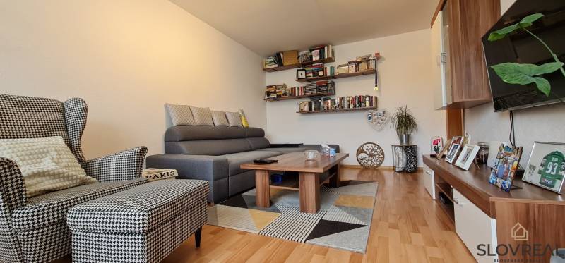 Predaj 2i byt v meste Kremnica, 64m2 - Obývačka v 2-izbovom byte s knihami, šedou sedačkou, kreslom a podlahou s dreveným dekorom.