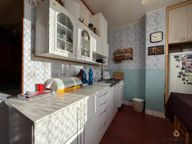 PREDAJ 2 IZBOVÝ BYT HANDLOVÁ  - Kuchyňa 2-izbového bytu so svetlou linkou, poličkou a retro hodinami na stene.