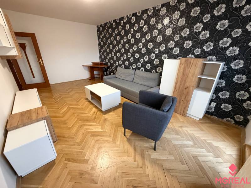 2 izbový byt s balkónom Topoľčany  - Obývačka v 3-izbovom byte s podlahou s dreveným dekorom, vzorovanou tapetou a nábytkom.