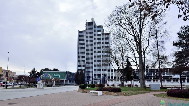 Kancelária s výhľadom na centrum Michaloviec. 16 m2 - Kancelárie na Námestí slobody v Michalovciach, vysoká budova s oknami a stromami v okolí.