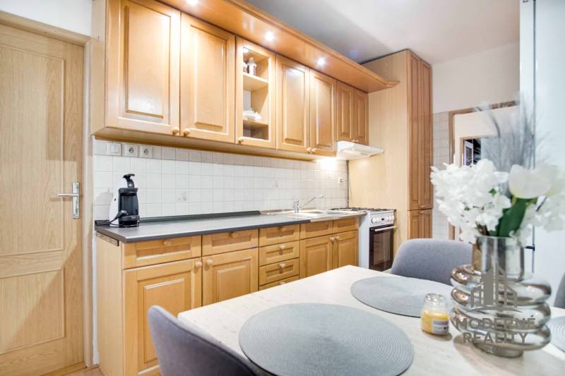 Na prenájom veľký 4-izbový byt s balkónom, garážou a záhradou, Kolta – - Kuchyňa v 4-izbovom byte s drevenými skrinkami, kvetmi a jedálenským stolom.