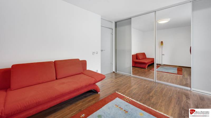 Novostavba 3 izbový byt, Palisády, 147 m2, výhľad, parkovanie, BA I - Izba v 3-izbovom byte s červeným gaučom, drevenou podlahou a zrkadlovou skriňou.