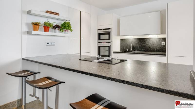 Novostavba 3 izbový byt, Palisády, 147 m2, výhľad, parkovanie, BA I - Kuchyňa v 3-izbovom byte s moderným ostrým a stoličkami, poličky s rastlinami.