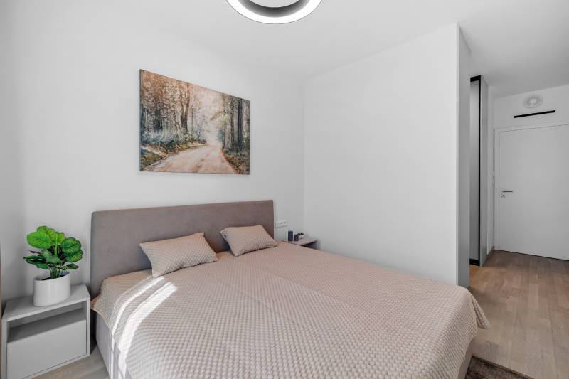 I Prenájom exkluzívneho 2-i bytu v projekte Metropolis - Spálňa v 2-izbovom byte s posteľou, obrazom a rastlinou, podlaha s dreveným dekorom.