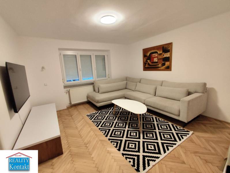 Na prenájom 2-izbový byt  Nové Zámky - Obývacia izba v 2-izbovom byte so sedačkou, televíziou a podlahou s dreveným dekorom.