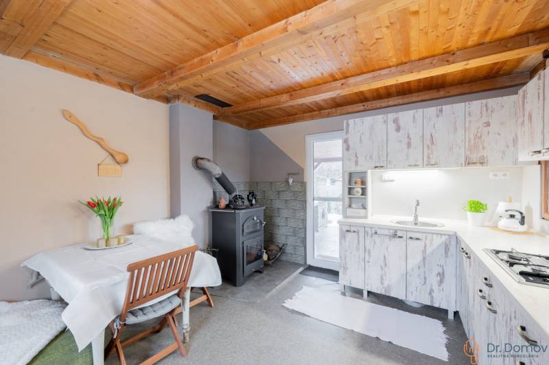 Záhradná chatka Handlová - Kuchyňa v chate s dreveným stropom, pecou a bielym nábytkom.